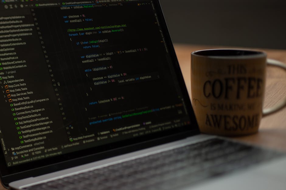 A close-up view of a laptop displaying lines of code on a dark screen, situated on a wooden desk. To the right of the laptop, there is a ceramic mug with a beige background and dark text that reads 'T